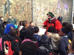 Image of Year 5 pupils, they are in the Great Hall at Hampton Court Palace listening to a talk about the Tudors by a member of staff dressed up in Tudor dress.
