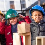 Image of Reception children wearing hard hats, using wooden 3d shapes to build different constructions.