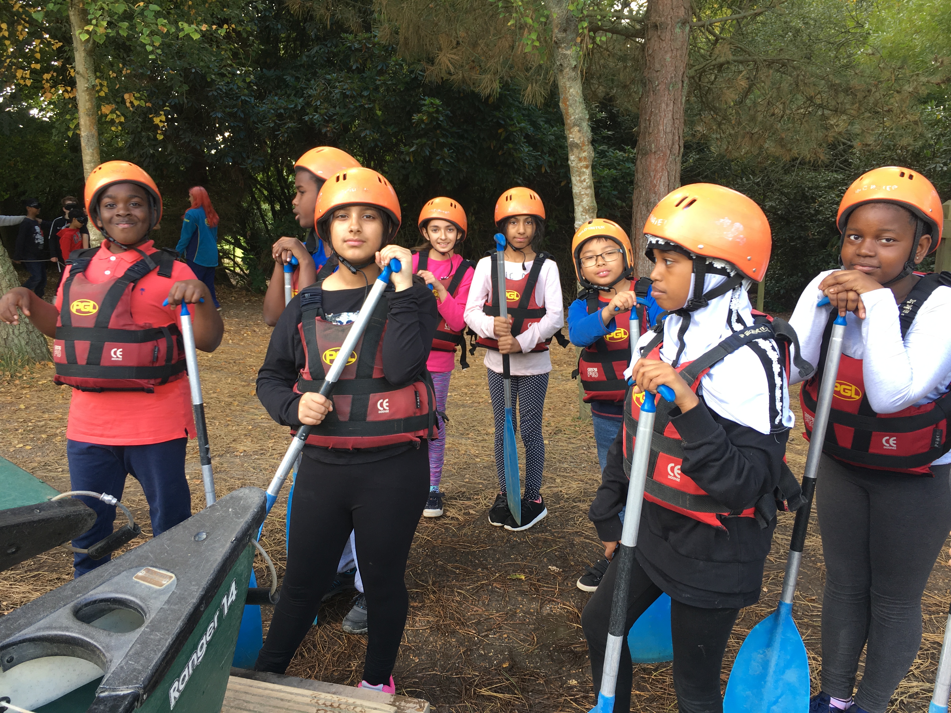 Image of a group of Year 6 children wearing hard hats and life jackets and standing next to canoes outside near a wooded area
