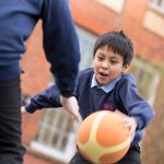 Image of A key stage 2 boy is holding a ball, playing basketball in the playground with a friend.
