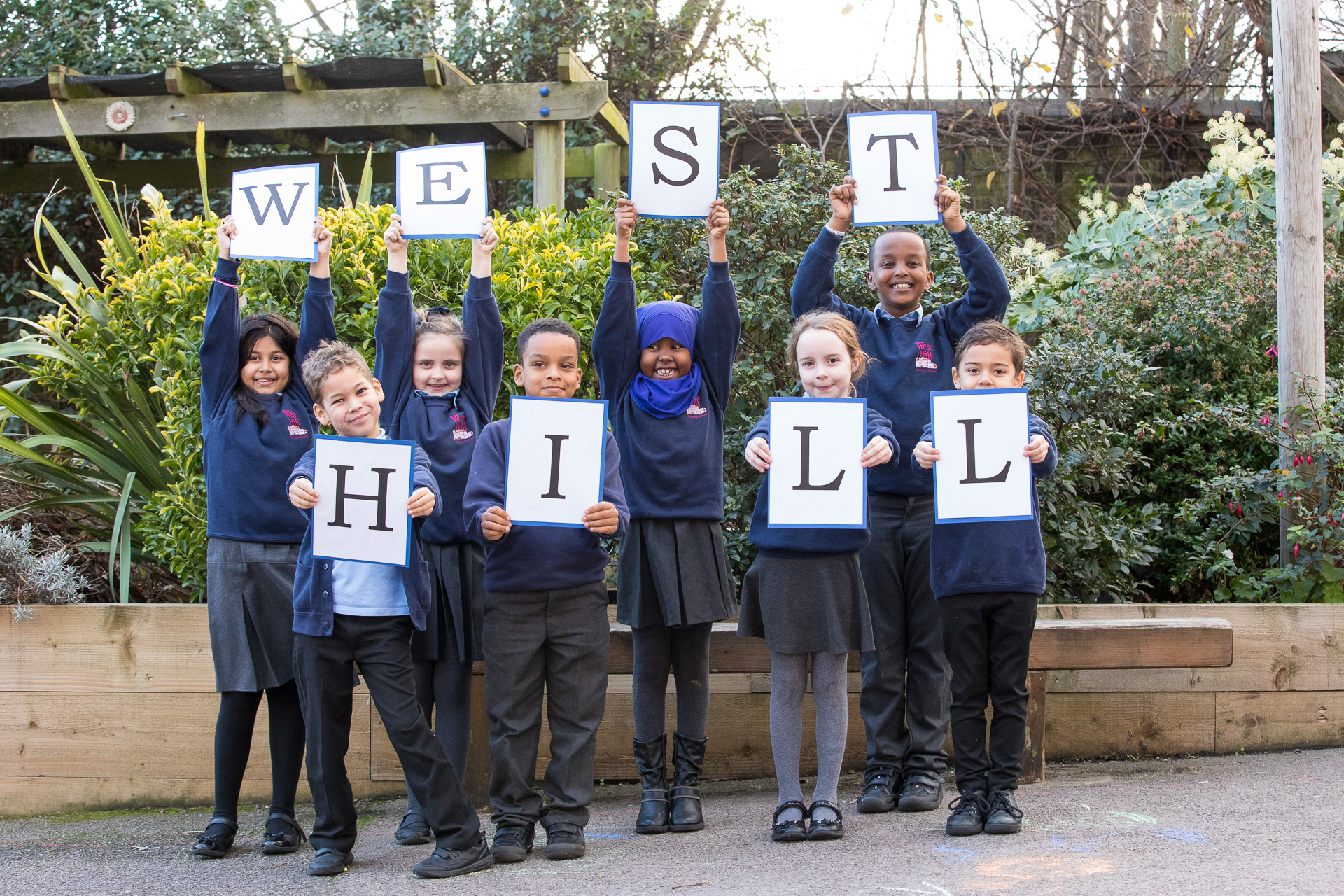 Image of eight pupils are standing in the playground in their school uniform each holding a large letter that together spell out the name West Hill.