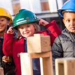 Image of Three Reception children wearing hard hats are outside and using wooden building blocks to make a construction.