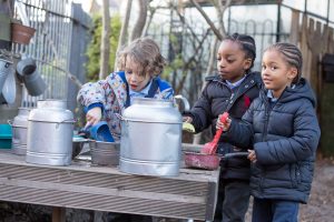 Image of three children are outside standing next to a table filling pans and large metal containers with mud.
