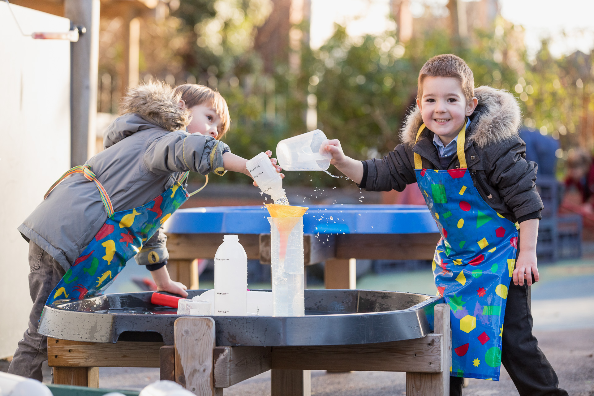 Image of two boys are outside wearing aprons, holding plastic jugs and playing with water.