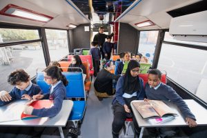 Image of A class of children are sitting at desks on the lower deck of a double decker bus looking at books.