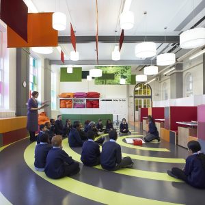 Image of A group of children are sitting on the floor of a brightly coloured hall with a teacher standing in front of them reading a book.