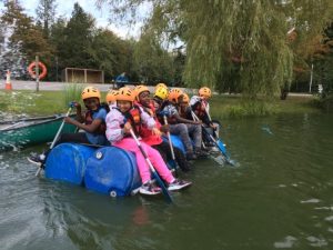 A group of children wearing life jackets and helmets are sitting on a raft, made out of plastic drums, on a small lake.